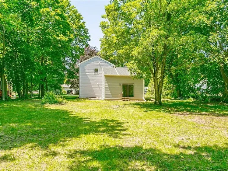 A house with white siding and a glass sliding door, surrounded by green grass and tall trees, under a bright blue sky.