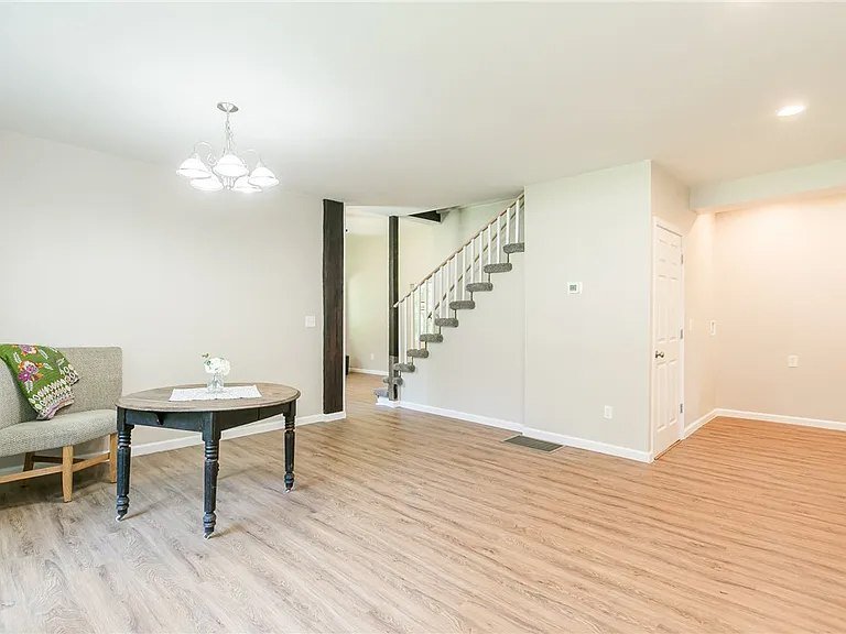 Empty living room with a small table, a bench with a colorful pillow, a staircase, and a white door.