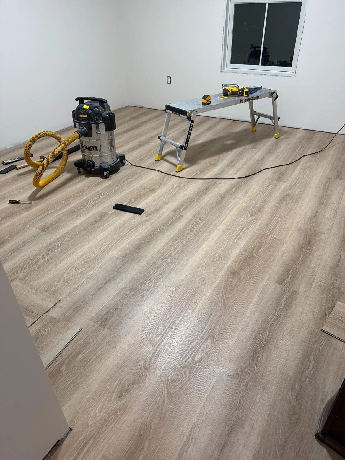 Room with new wood laminate flooring, work tools including a table saw, vacuum, and various small tools, and a window on the wall.