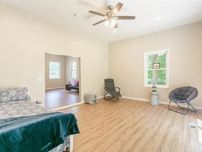 A spacious, minimally furnished living room with two chairs, a small side table, and a ceiling fan. The room has light-colored walls, wooden flooring, and one window showing green trees outside.