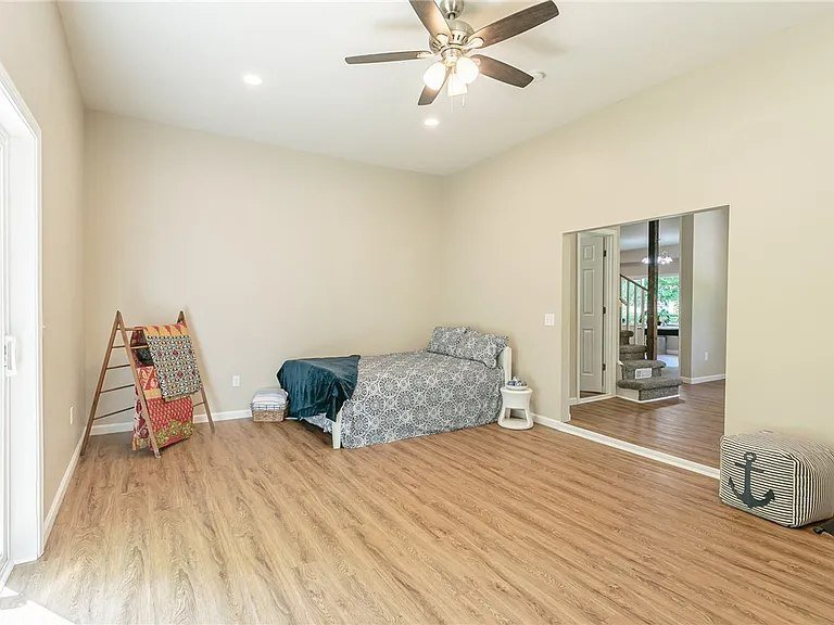 A mostly empty living room with light-colored walls, a ceiling fan, and wooden flooring. There is a twin bed with a patterned comforter, a small round white table next to it, a folded blanket on the bed, and a decorative item with an anchor design on