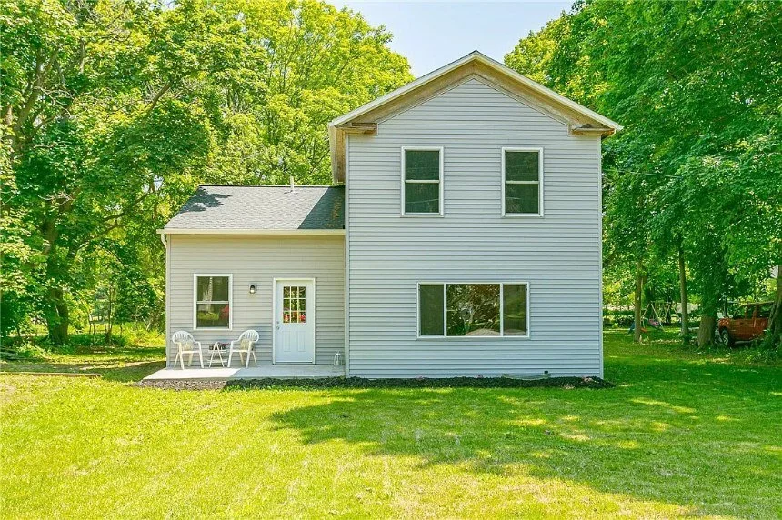 A two-story house with light gray siding, surrounded by green trees and grass, featuring a small porch with two white chairs and a door, and multiple windows.
