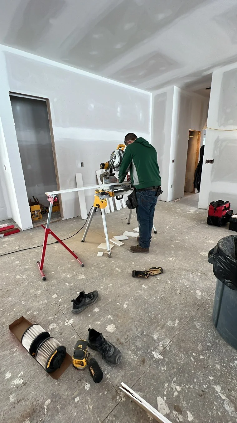 A person working with a saw on a construction site inside a room with unfinished drywall and open doorways. Various tools and shoes are scattered on the floor.