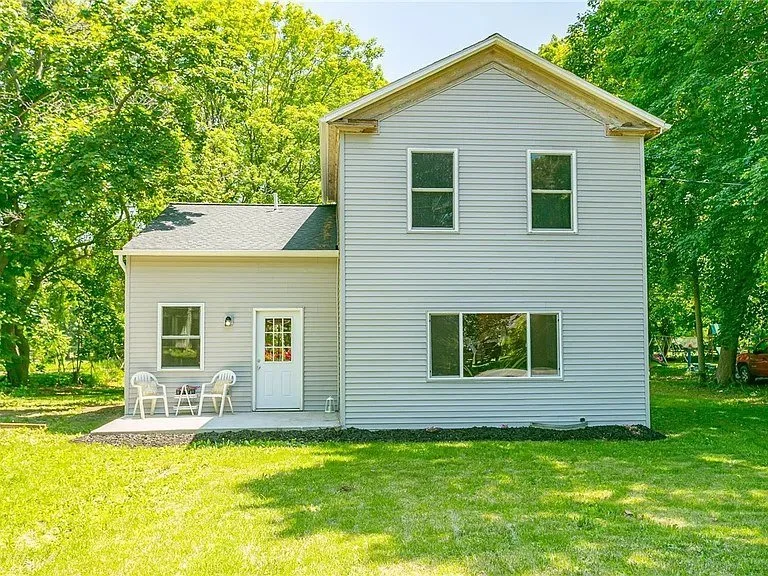 A two-story house with white siding, several windows, and a small back porch with white plastic chairs, surrounded by green trees and lawn.
