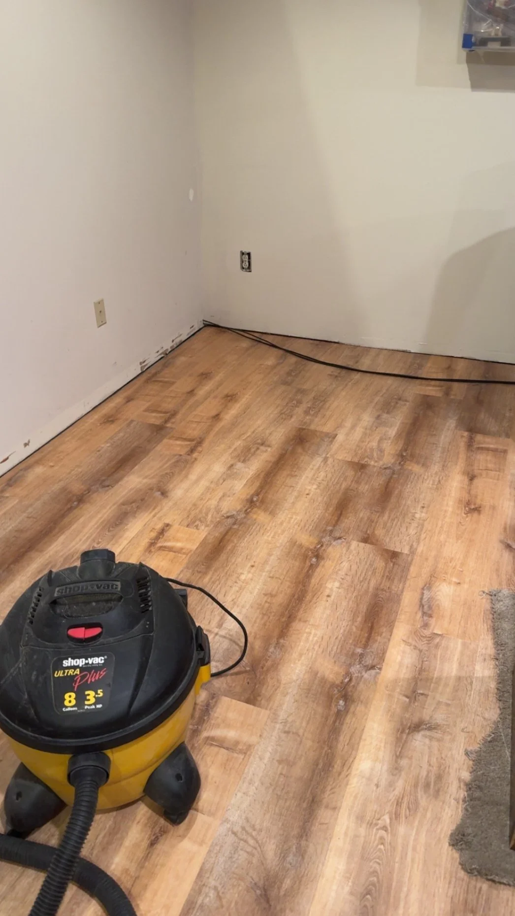 A yellow Shop-Vac vacuum cleaner on a wooden floor in a room with white walls and electrical outlets, part of a flooring renovation.