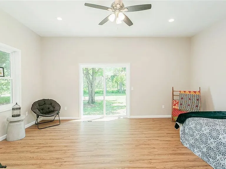 Empty living room with white walls, hardwood floor, ceiling fan, sliding glass door to outdoor view, a chair, a small side table with a decorative birdcage, and a bed with a quilted blanket.