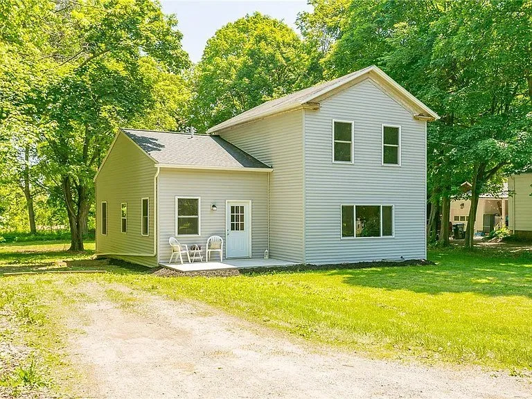 Two-story house with light gray siding, white trim, and a small porch with two plastic chairs, situated in a grassy yard with large trees.