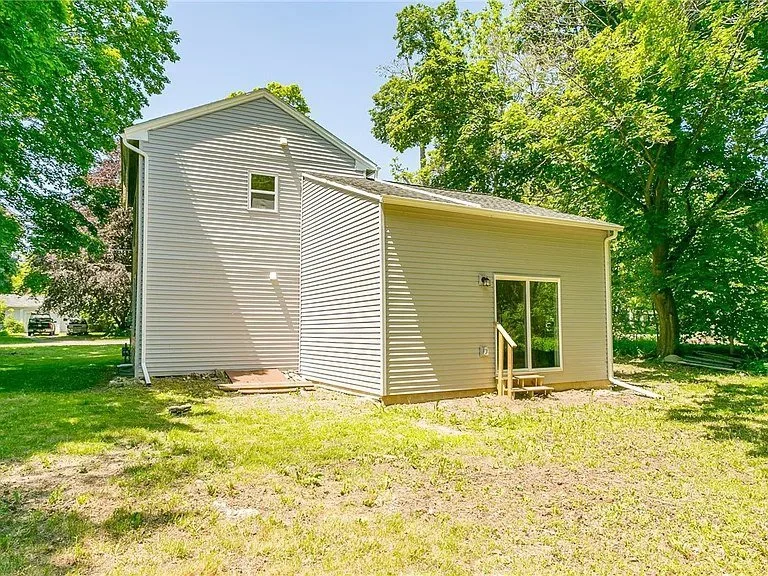 Back of a two-story house with beige siding, sliding glass door, small backyard with green grass, and surrounded by tall trees.