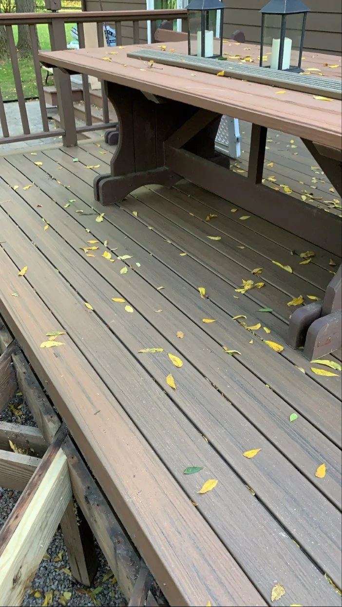 A wooden deck with a wooden picnic table on it, scattered with yellow leaves and a few green leaves, with lanterns on the table.