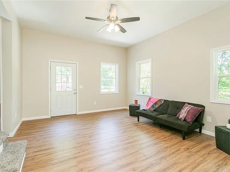 Empty living room with beige walls, wood flooring, three windows, a black sofa with pillows and a blanket, two side tables, ceiling fan, and a door.