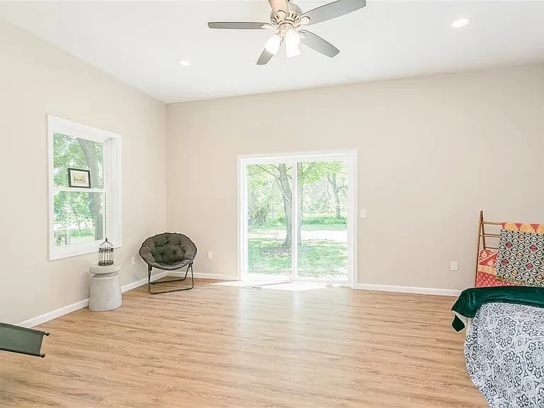 Empty living room with light-colored walls, a ceiling fan, a window, a sliding door, a folding chair, and a bed with a quilted headboard.