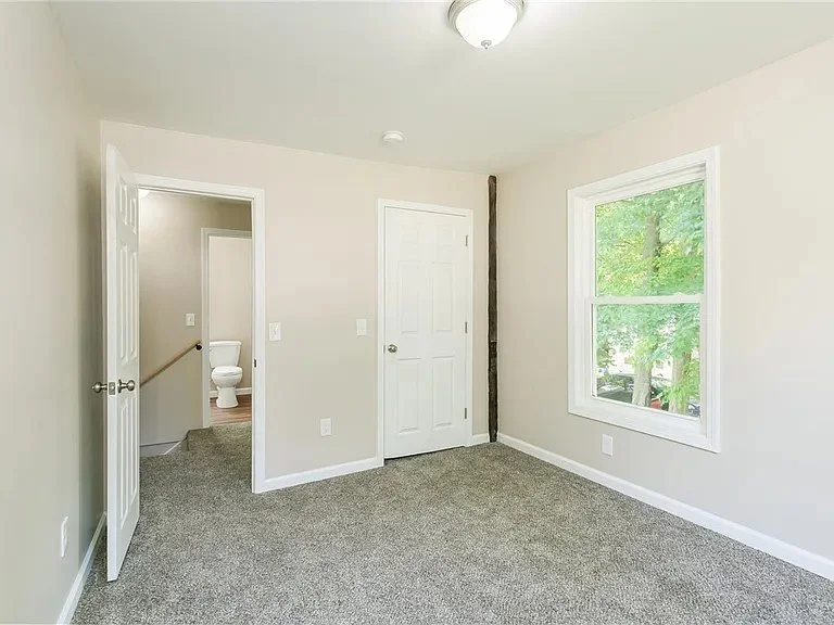 Empty bedroom with beige walls, a window showing green trees outside, and a bathroom door slightly open revealing a toilet.
