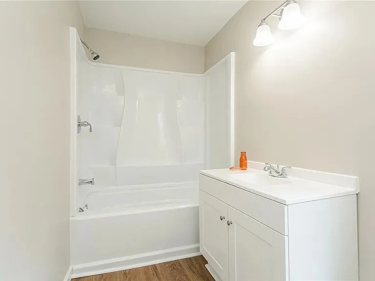 Bathroom with white bathtub shower combo, white vanity with sink, mirror with three light fixtures, and beige walls.