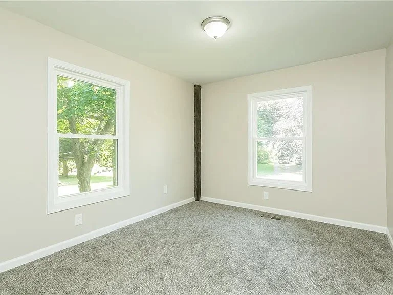 Empty room with beige walls, two windows, gray carpet, and ceiling light fixture.