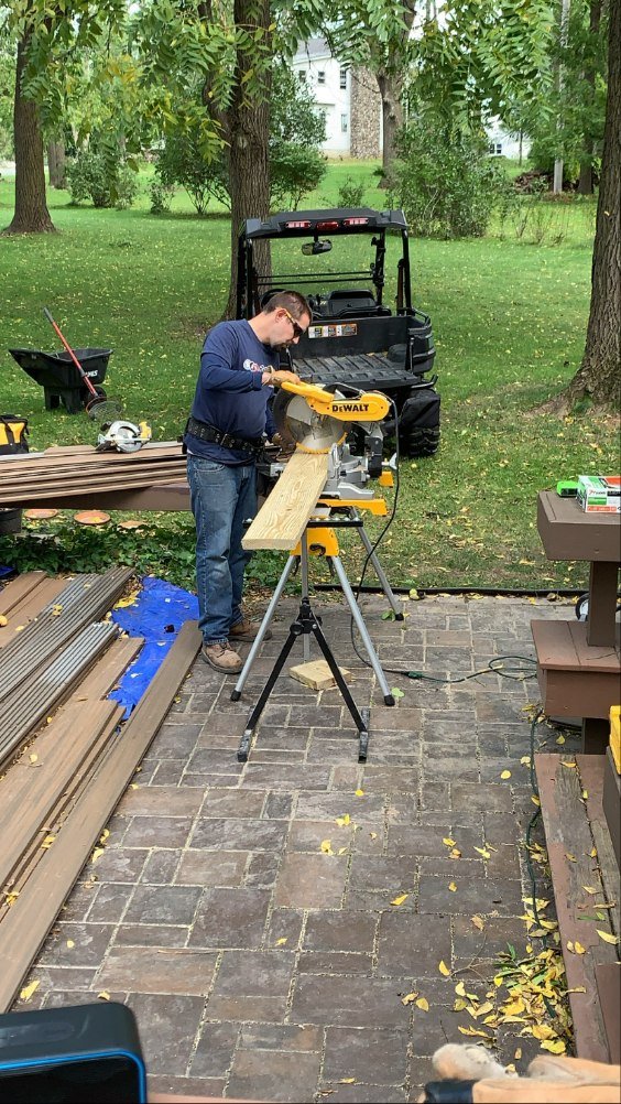 A man is using a miter saw to cut a piece of wood outdoors in a backyard, with trees and grass in the background.
