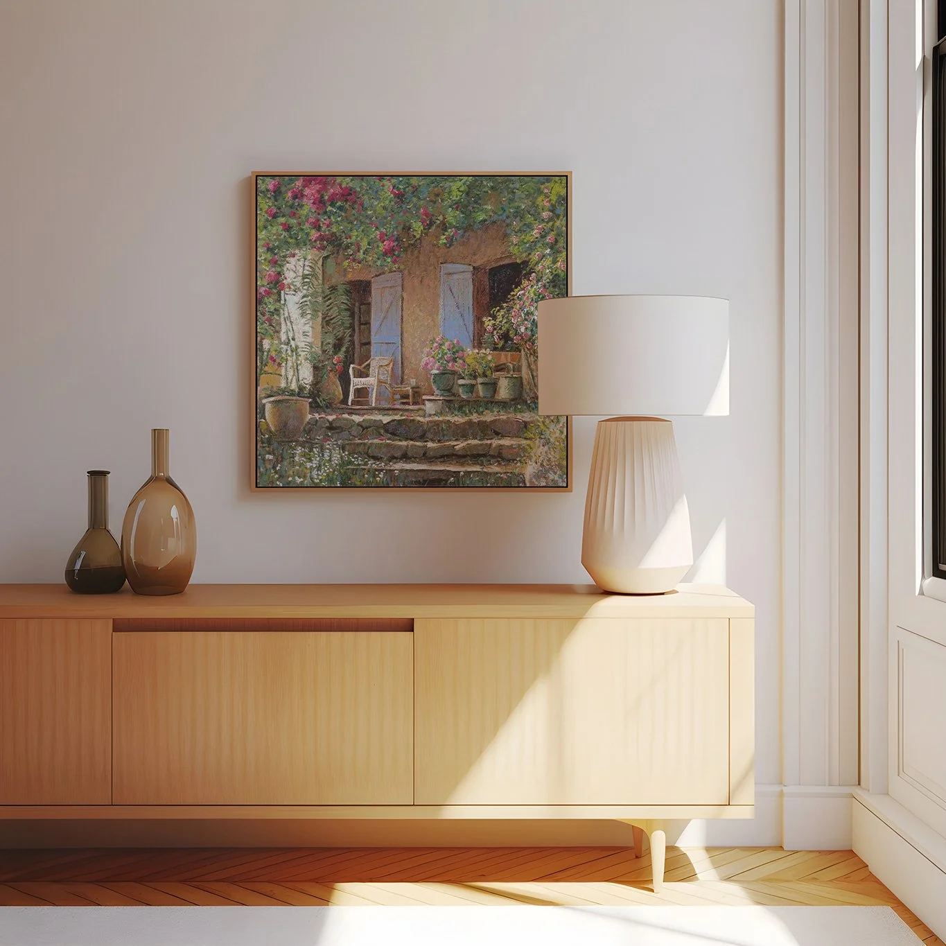 Decoração de sala com quadro decorativo de um jardim em uma parede branca, luminária de mesa e dois vasos na mobília de madeira clara, com luz natural entrando pela janela.