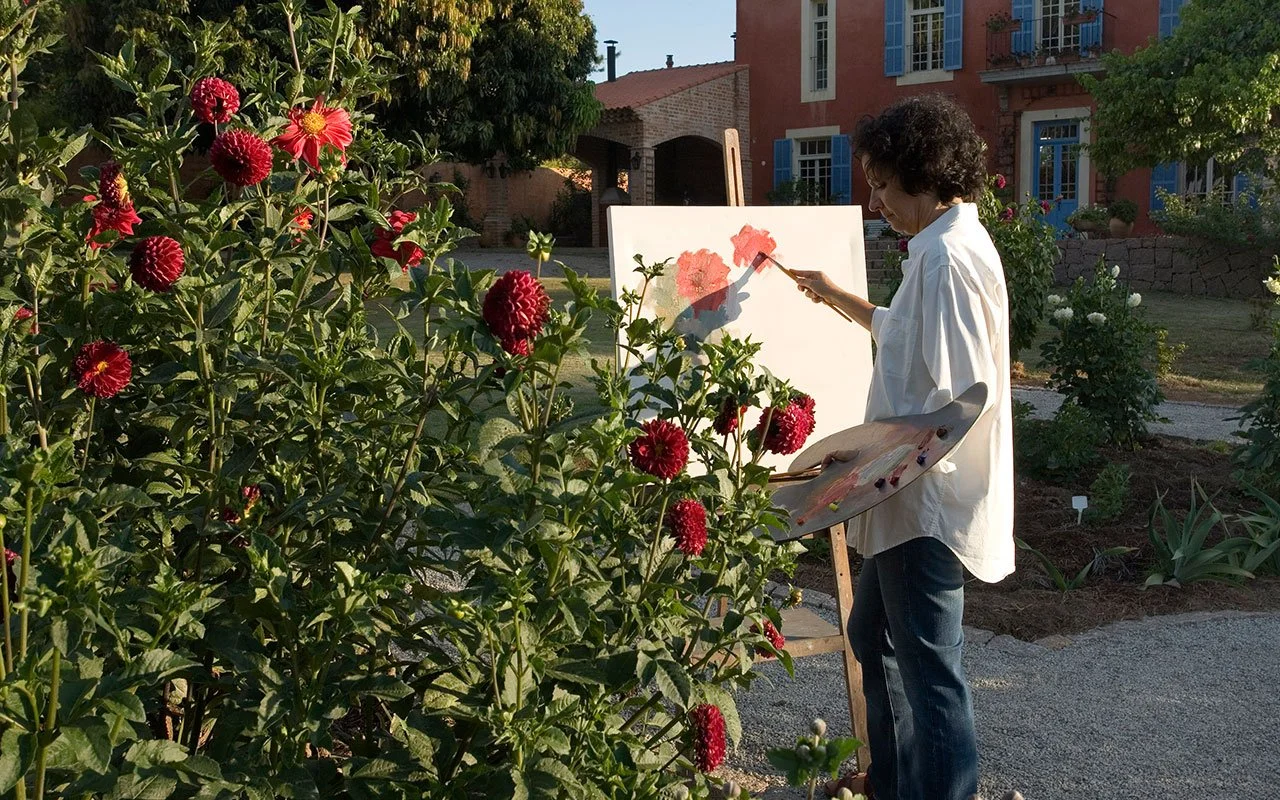 Pessoa pintando uma paisagem de um jardim com várias plantas e flores, usando uma tela e pincéis ao ar livre.