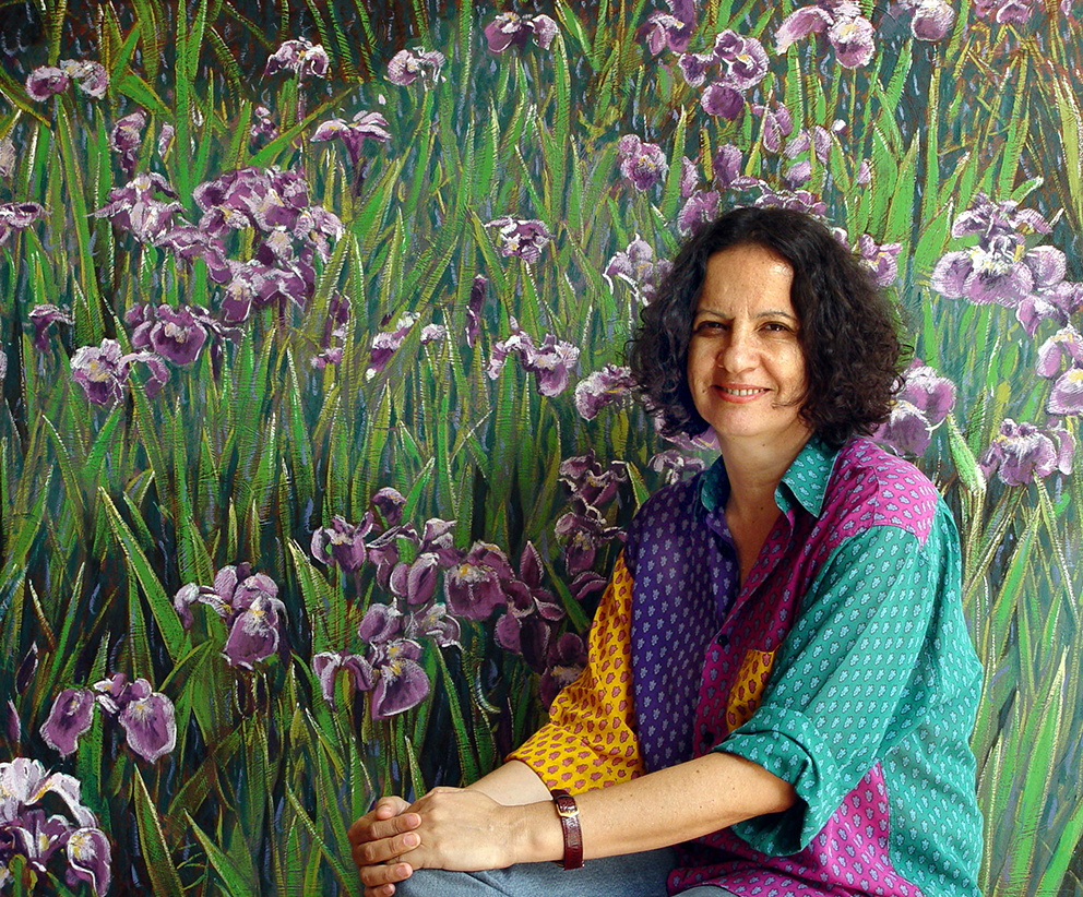 Mulher sorridente sentada na frente de um fundo de pintura que representa um campo de flores roxas.