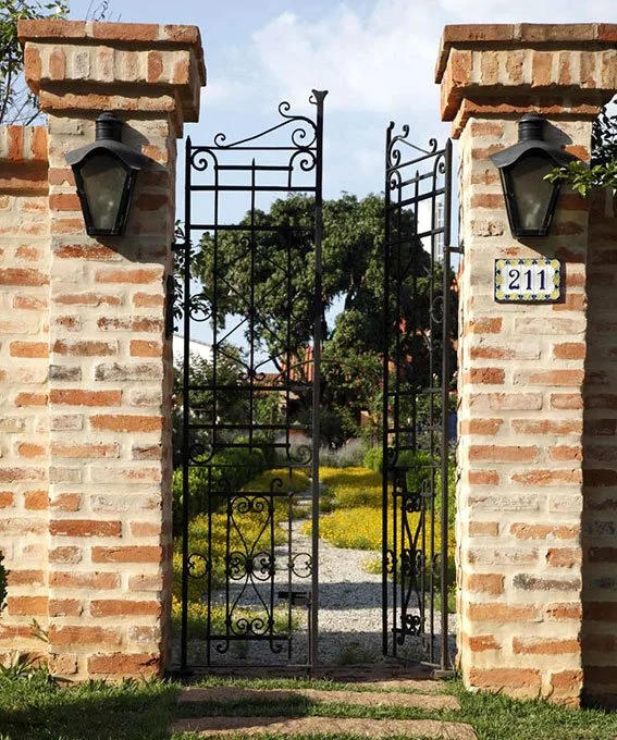 Portão de ferro preto entre duas colunas de tijolos com luminárias nas laterais e endereço 211 em um azulejo nas colunas, levando a um jardim com árvores e caminho de pedra.