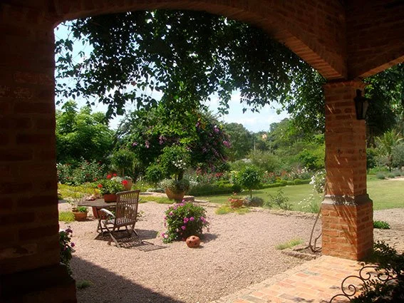 Visão de um jardim com móveis de jardim, plantas, árvores e flores, visto de uma varanda com pilares de tijolos.