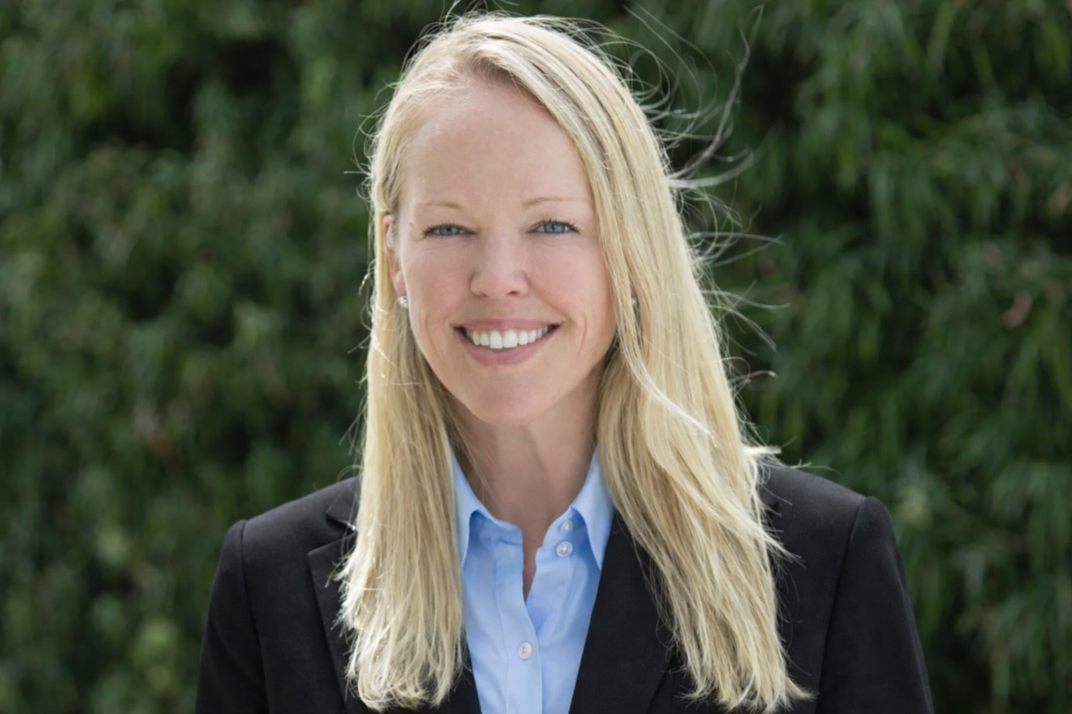 A woman, Max Powers, with long blonde hair, wearing a black blazer and a light blue shirt, smiling outdoors against a green leafy background.