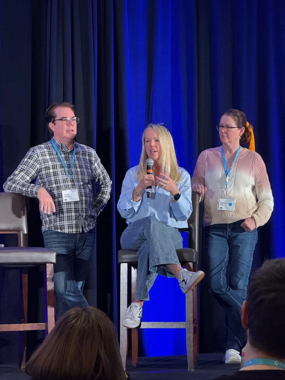Max P on stage participating in a panel discussion, seated on a stool, and two others standing on either side, all wearing lanyards with name badges, set against a dark curtain background with blue lighting. Max Powers presenting to an organization.