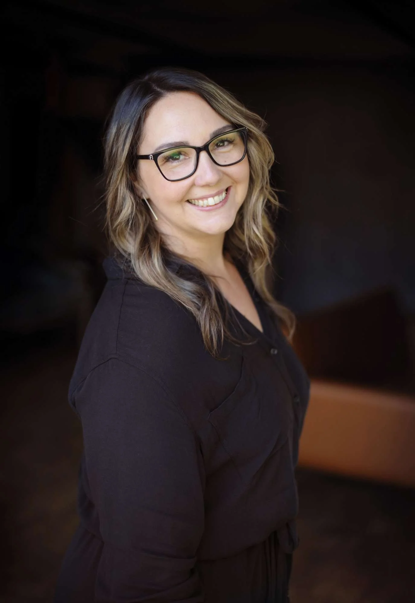 Jenna Johnson, owner of Avenue 530 Realty, wearing glasses and wavy hair smiling in front of a dark blurred background.