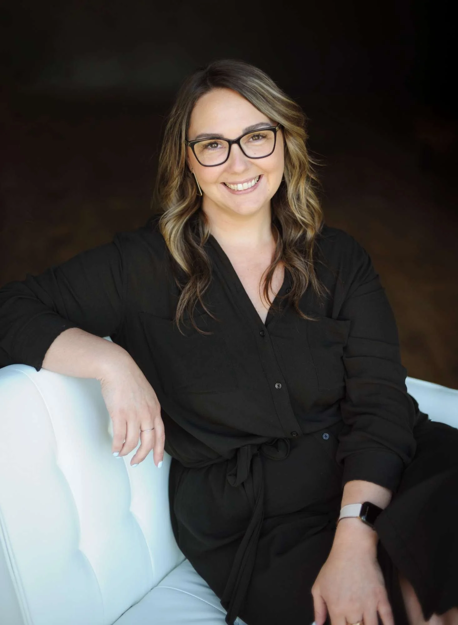 Jenna Johnson, owner of Avenue 530 Realty, with wavy brown hair, glasses, and a black shirt sitting on a white chair. She is smiling and looking at the camera, with her left arm resting on the chair's back and her right arm on her lap.