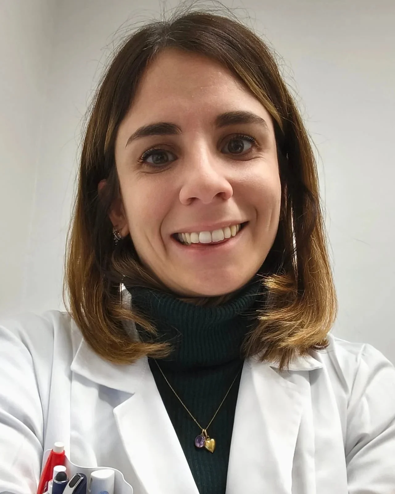 Donna con capelli castani e occhi castani, sorridente, indossa una giacca bianca, una maglia nera a collo alto, una collana con pendente a cuore. Sul tavolo ci sono penne e marker.