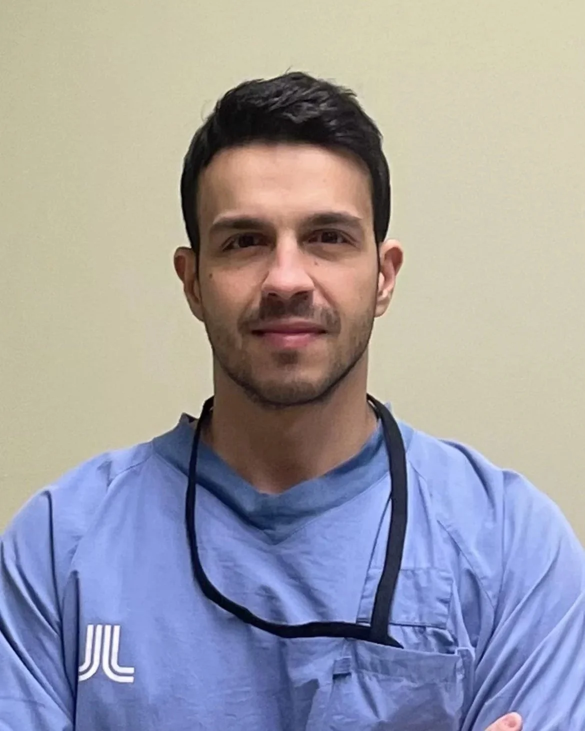 Un uomo con capelli scuri, vestito con un camice blu, sorridente, in un ambiente chiaro.
