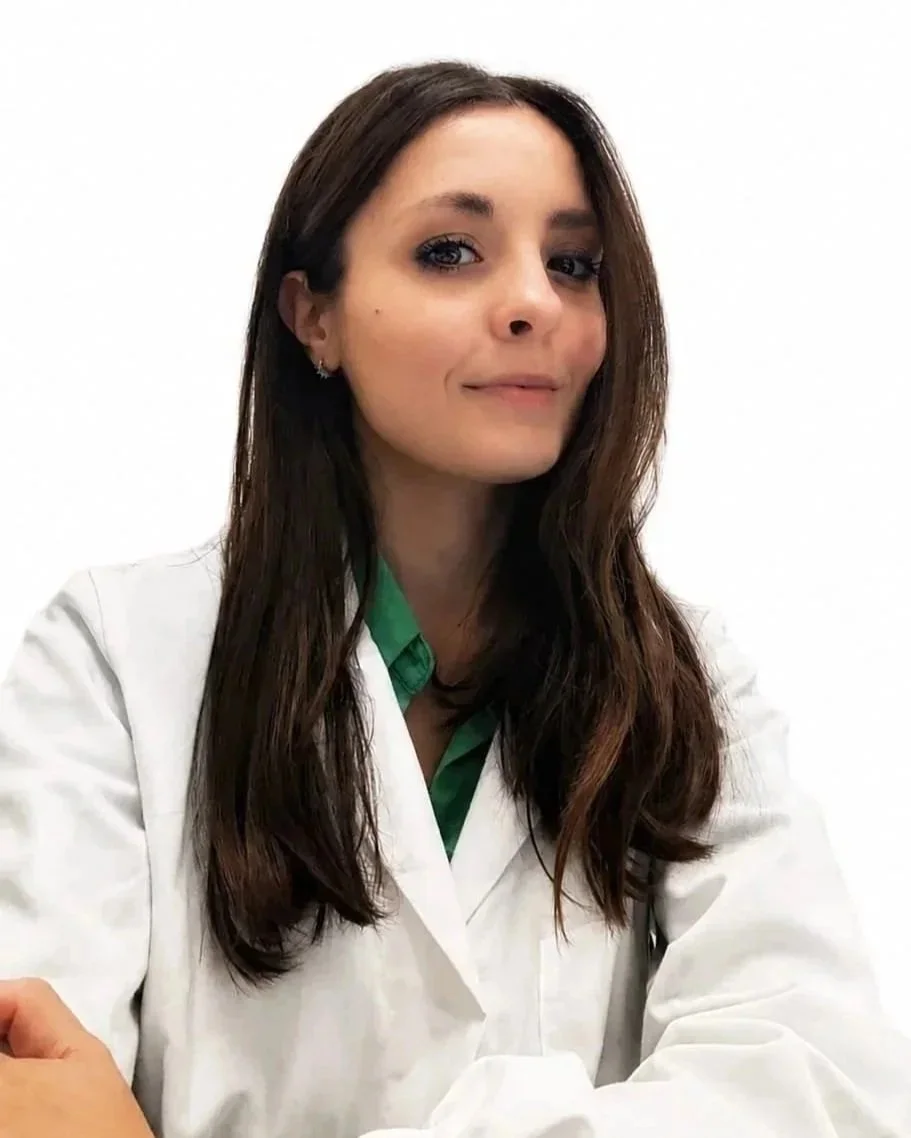 Donna con capelli castani lunghi, occhi azzurri, indossa una camicia verde e un camice bianco, sorridente, sfondo bianco.