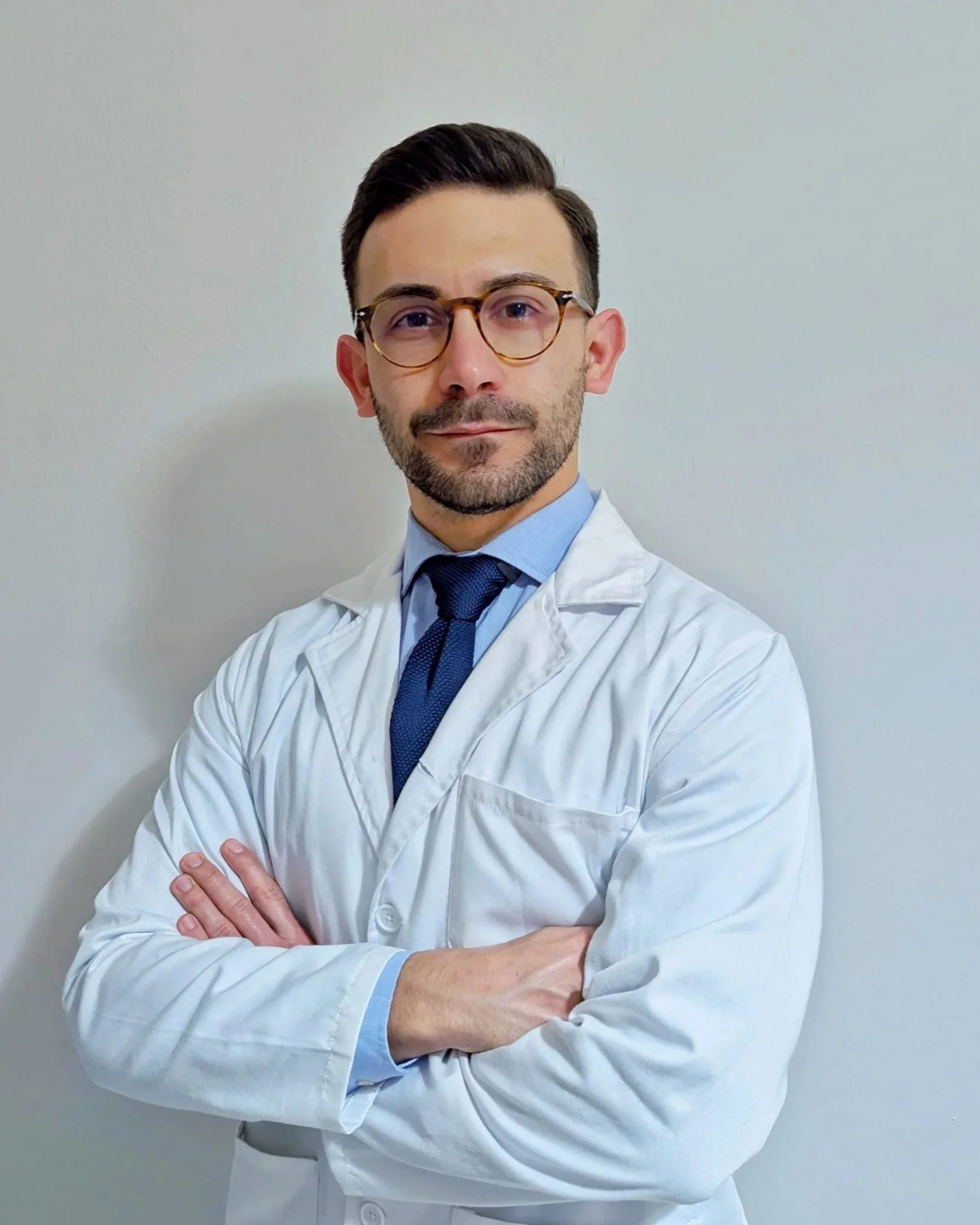 Un uomo medico con occhiali e barba, indossa un camice bianco e una camicia blu con cravatta blu scuro, si trova contro uno sfondo grigio, posa con le braccia incrociate.