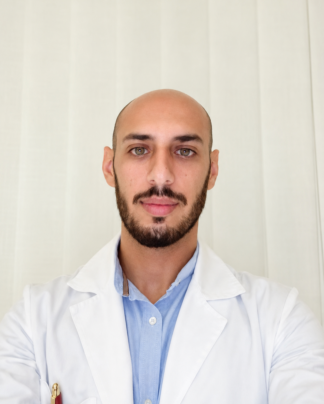 Un uomo con capelli rasati, barba e camice bianco si scatta un selfie di fronte a uno sfondo chiaro.