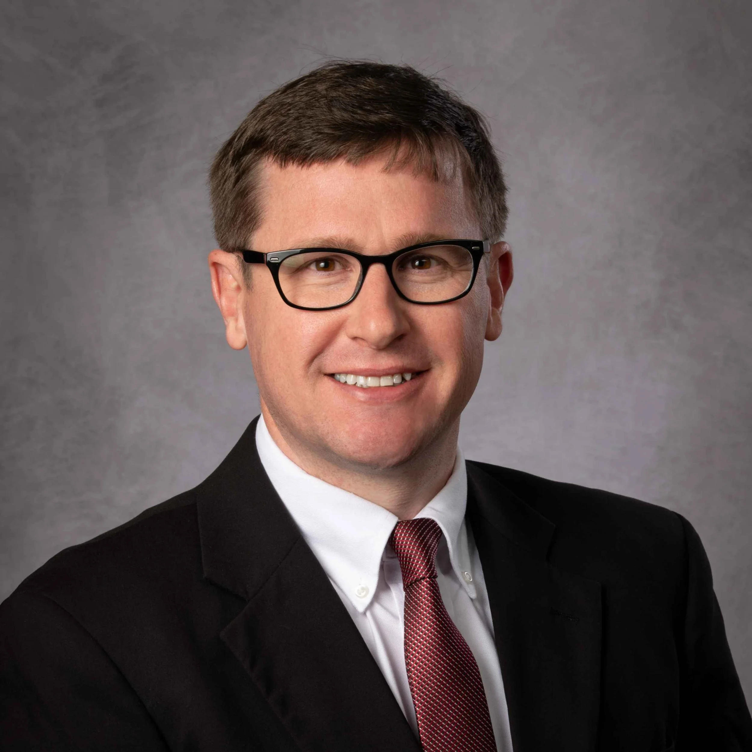 Close-up of a man wearing glasses, in a suit and tie, smiling against a gray background.