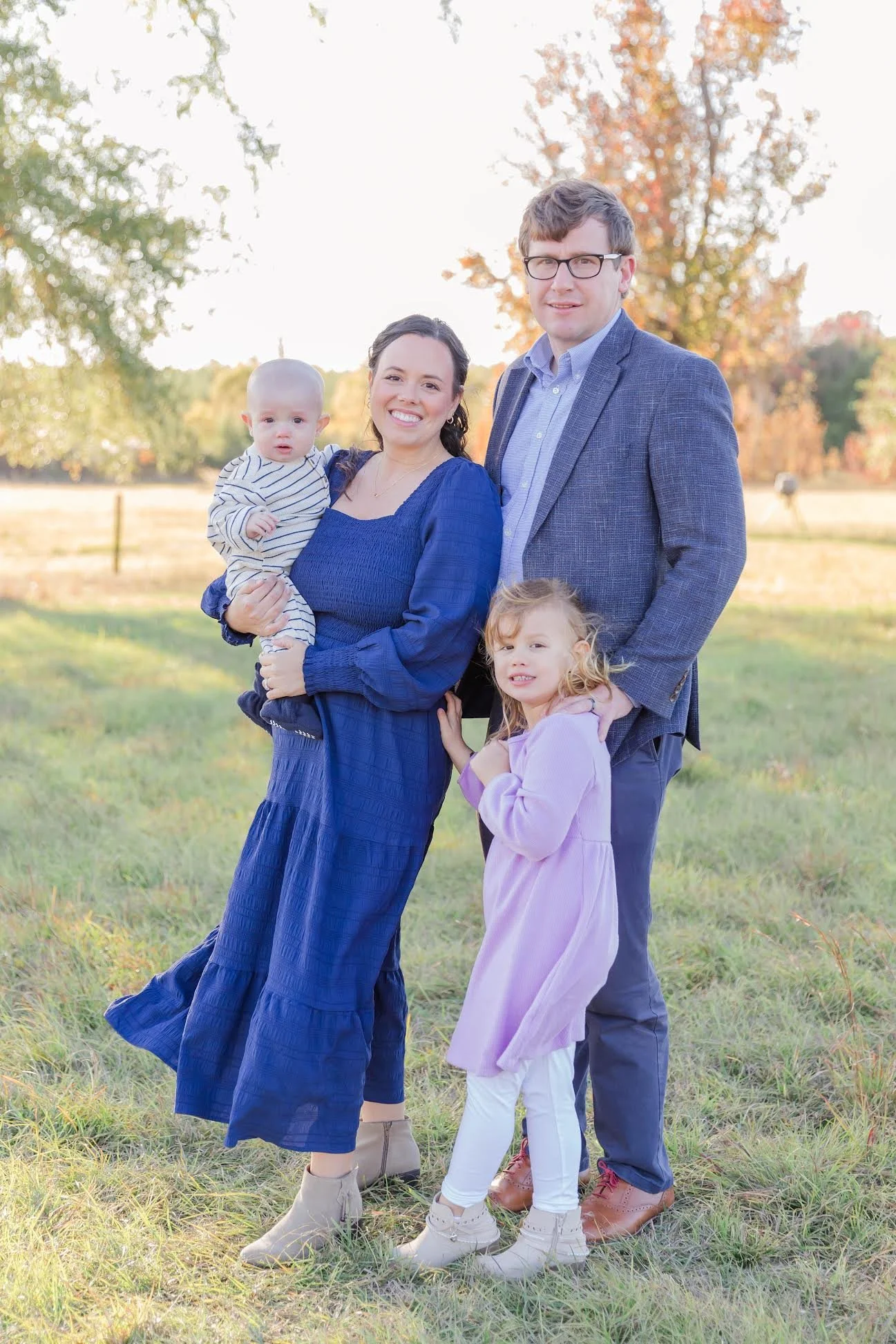 A family of four standing outdoors in a field with trees in the background during fall, smiling at the camera. The mother holds a baby, and a young girl is standing next to the father. All are dressed in casual, colorful clothing.