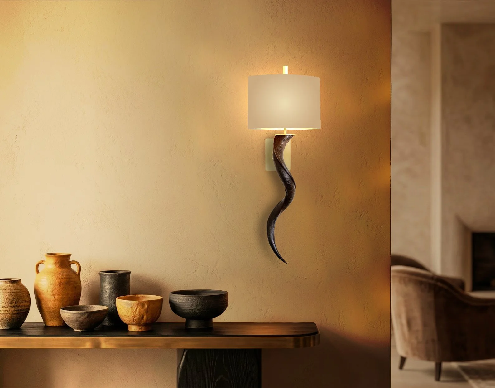 A beige wall with a modern wall-mounted sconce light. Below, a wooden table holds various ceramic pots and bowls in earthy tones. To the right, part of a beige armchair with dark wooden legs is visible in the background.