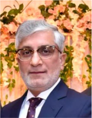 A man with gray hair, glasses, wearing a dark suit and tie, standing in front of a floral backdrop.