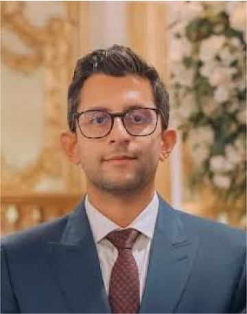 A man wearing glasses in a suit and tie standing in an ornate decorated room with floral arrangements in the background.