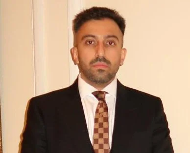A man in a black suit and brown checkered tie standing indoors against a plain wall.