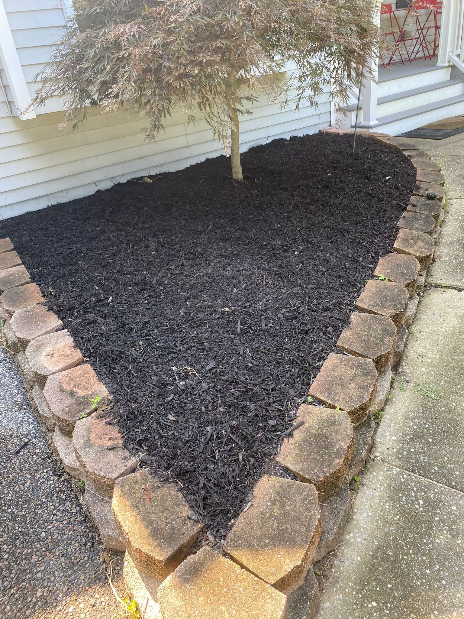 A flower bed with dark mulch surrounded by irregularly shaped brick border, located next to a light-colored house with horizontal siding and an outside staircase with red chairs visible on the porch.