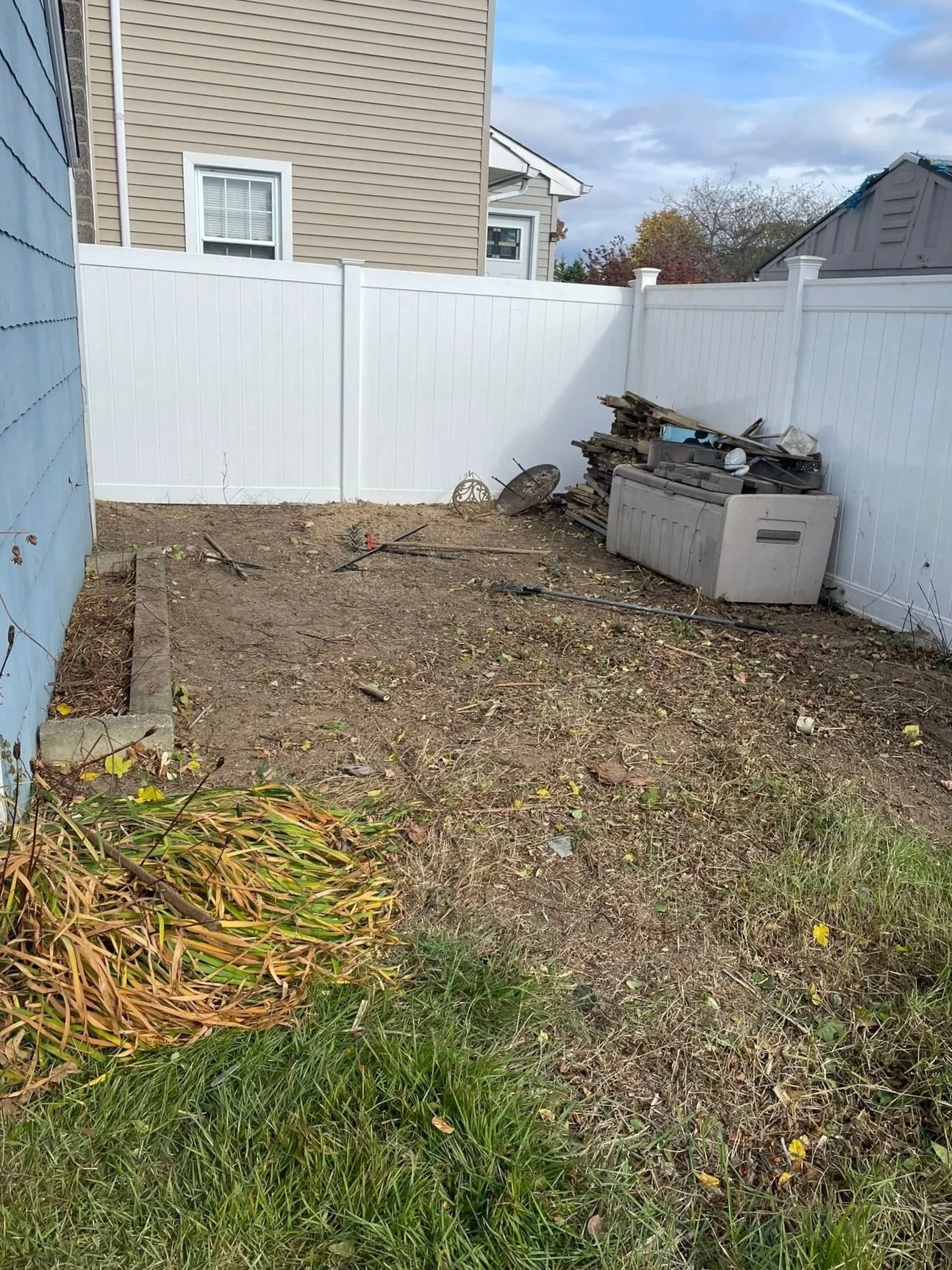 Empty backyard with dirt ground, a pile of wood and metal scrap, a garden hose, and some plants and grass.