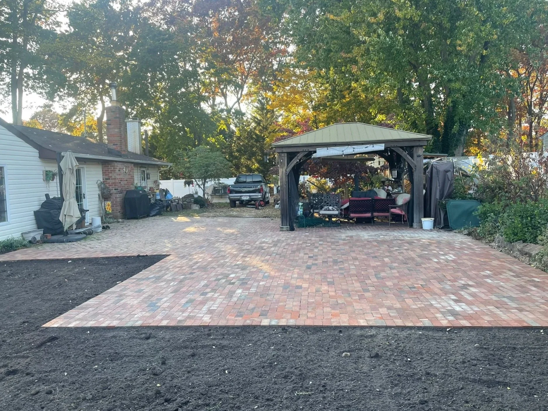 Backyard with a brick patio, a covered seating area with outdoor furniture, a parked truck, a brick chimney on the house, and trees with autumn foliage in the background.