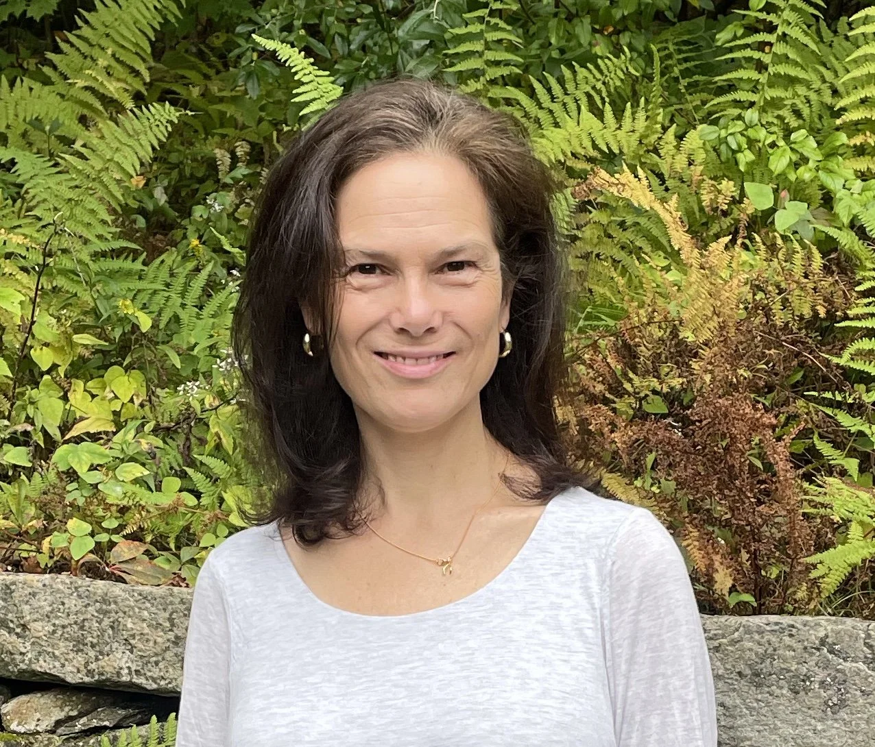 A woman smiling outdoors with a background of lush green ferns and plants, wearing a white top, pearl earrings, and a delicate necklace.