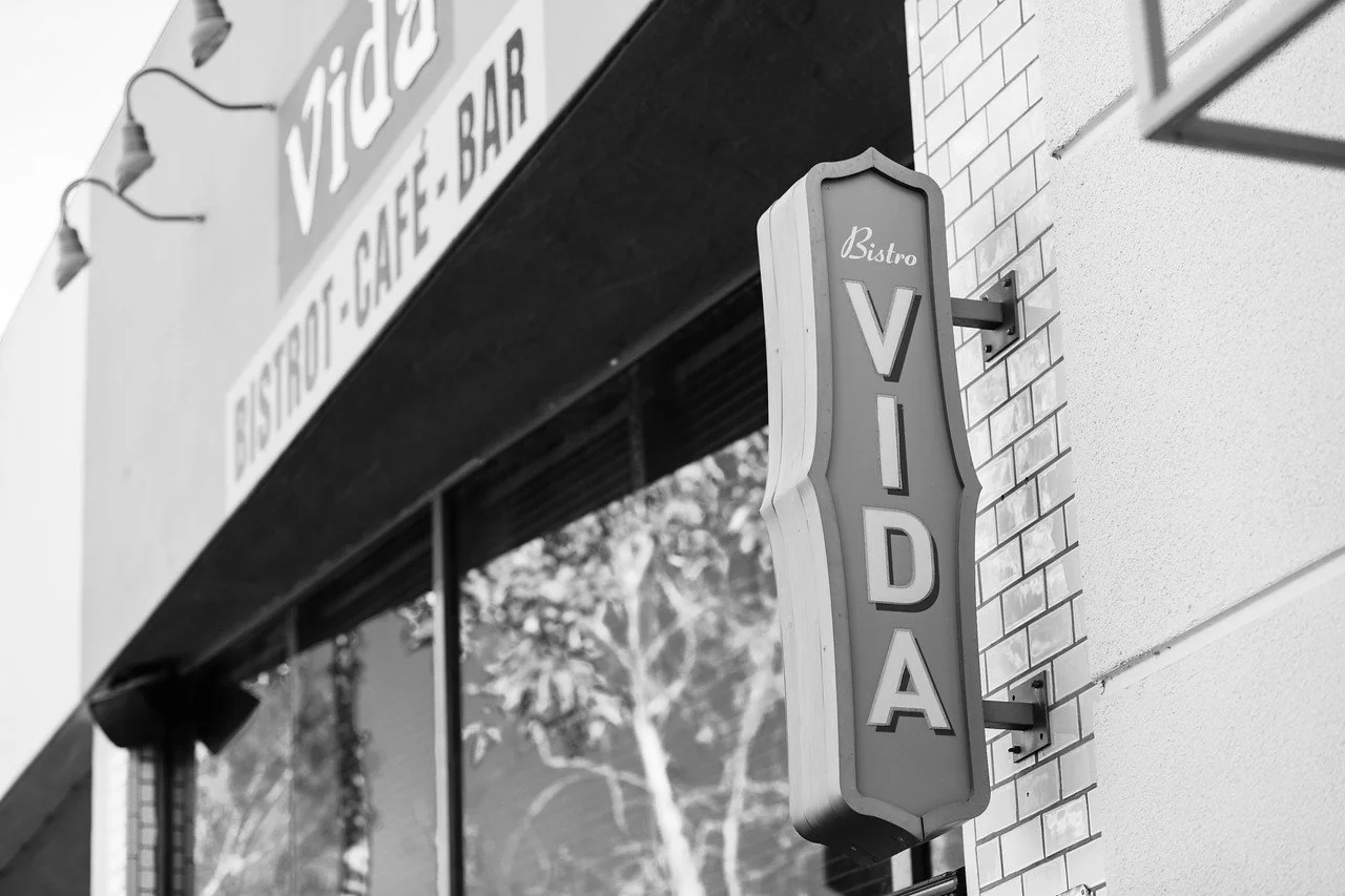 Black and white photo of a vertical sign that reads Bistro Vida on the side of a brick building, with a window reflecting trees in the background.