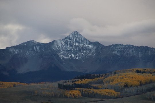 A snow-capped mountain with a forest and rolling hills in the foreground under a cloudy sky.