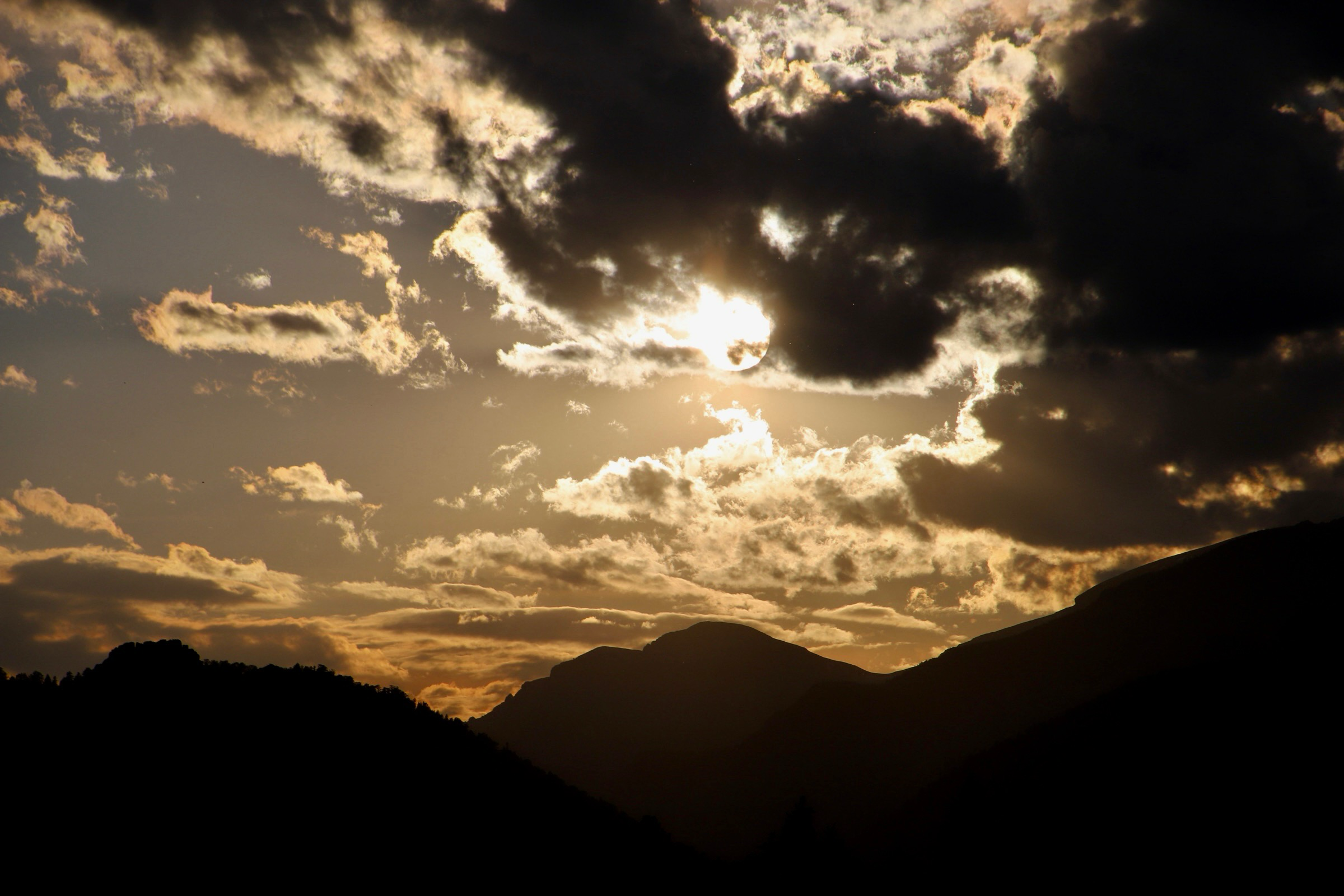Sun partially hidden by dark clouds over silhouettes of mountains and trees at sunset.
