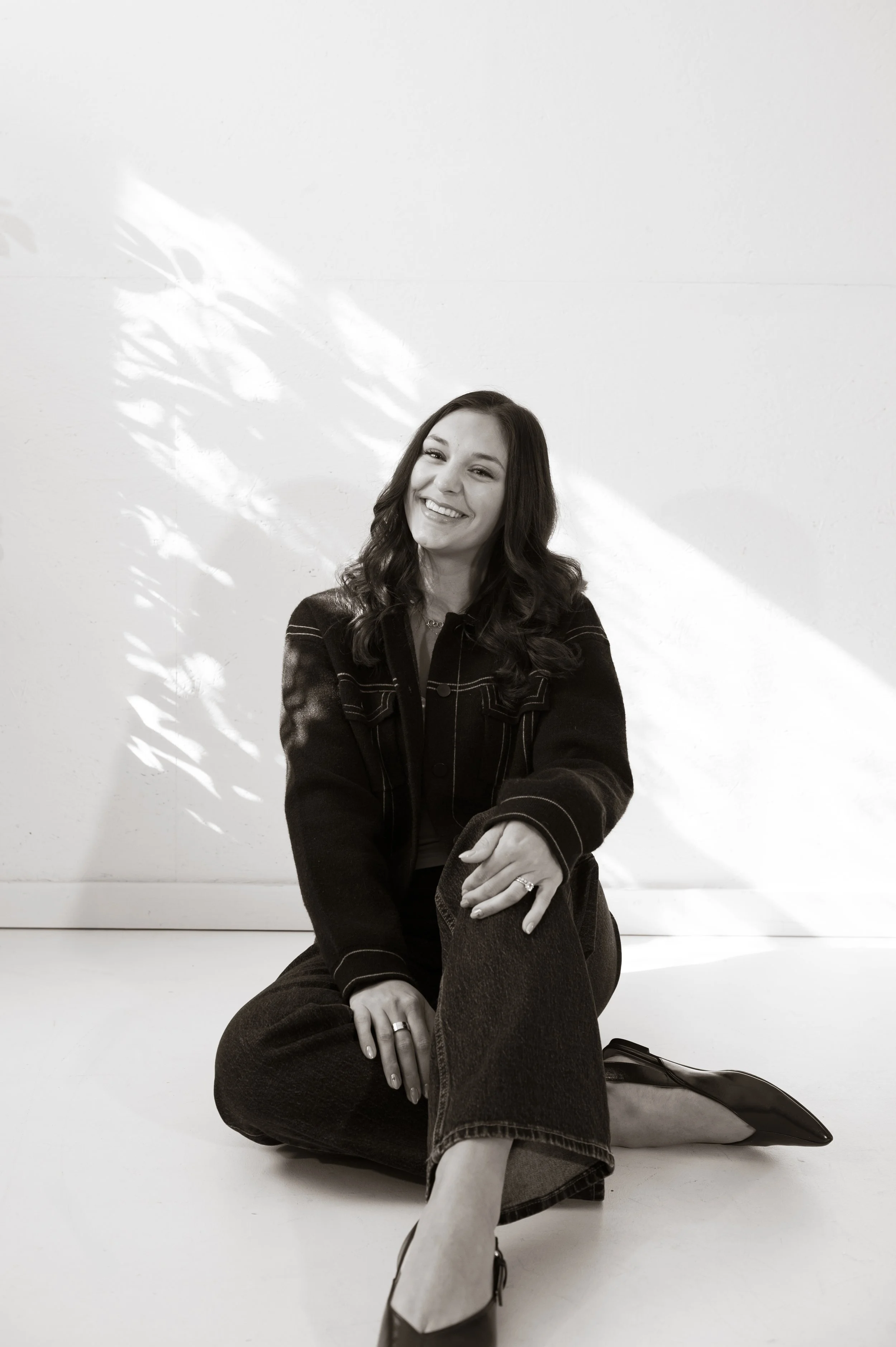 Black and white photo of a woman sitting cross-legged on the floor, smiling, with a plain wall in the background, light shadows, and a backpack beside her.