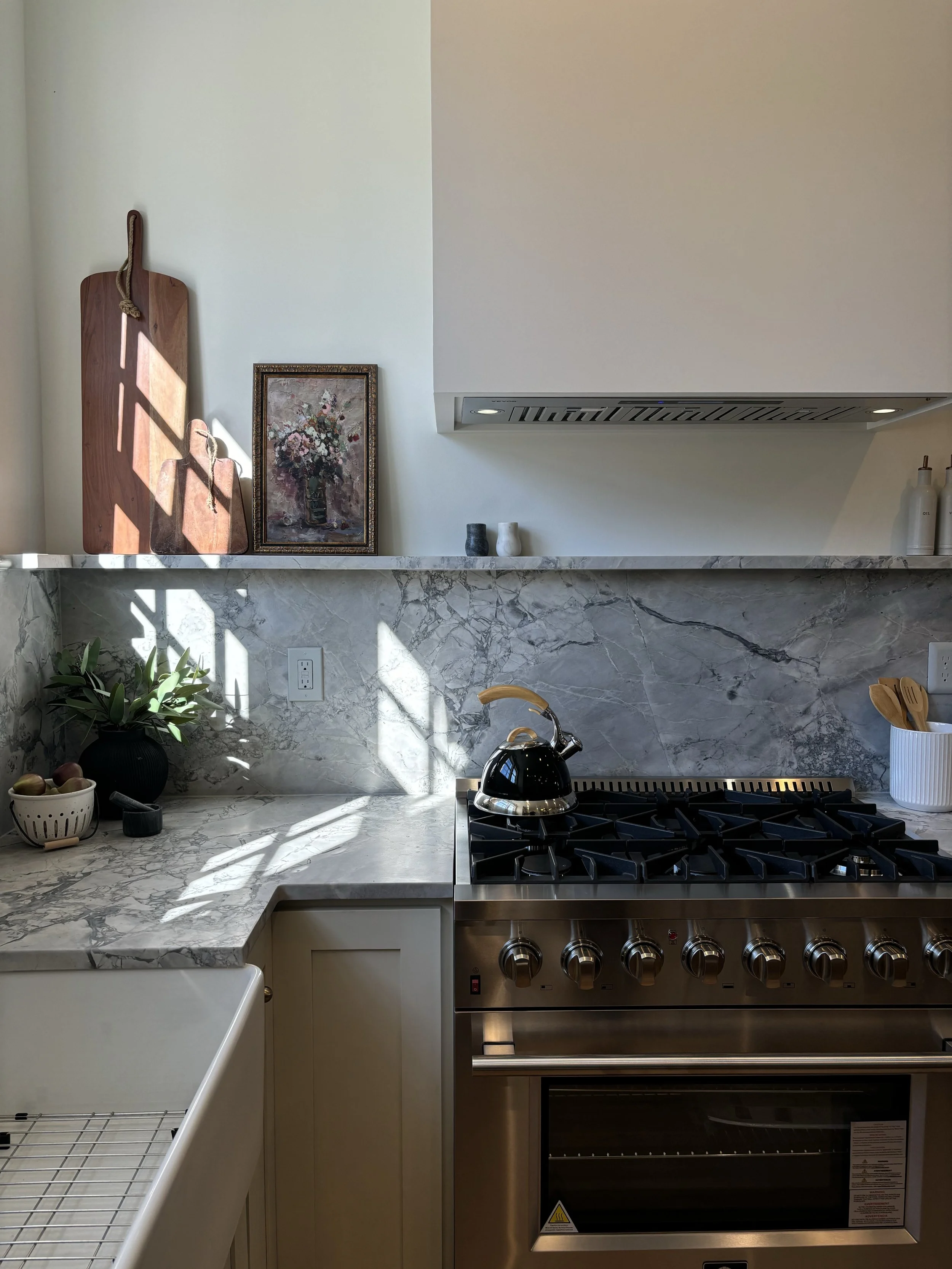 A kitchen with a marble countertop, a stainless steel stove, and a white range hood. On the countertop are a framed floral painting, two vases, and bulbs of garlic. To the left, there is a black plant, and next to it, a small bowl of apples. There is a white dish drying rack in the foreground.