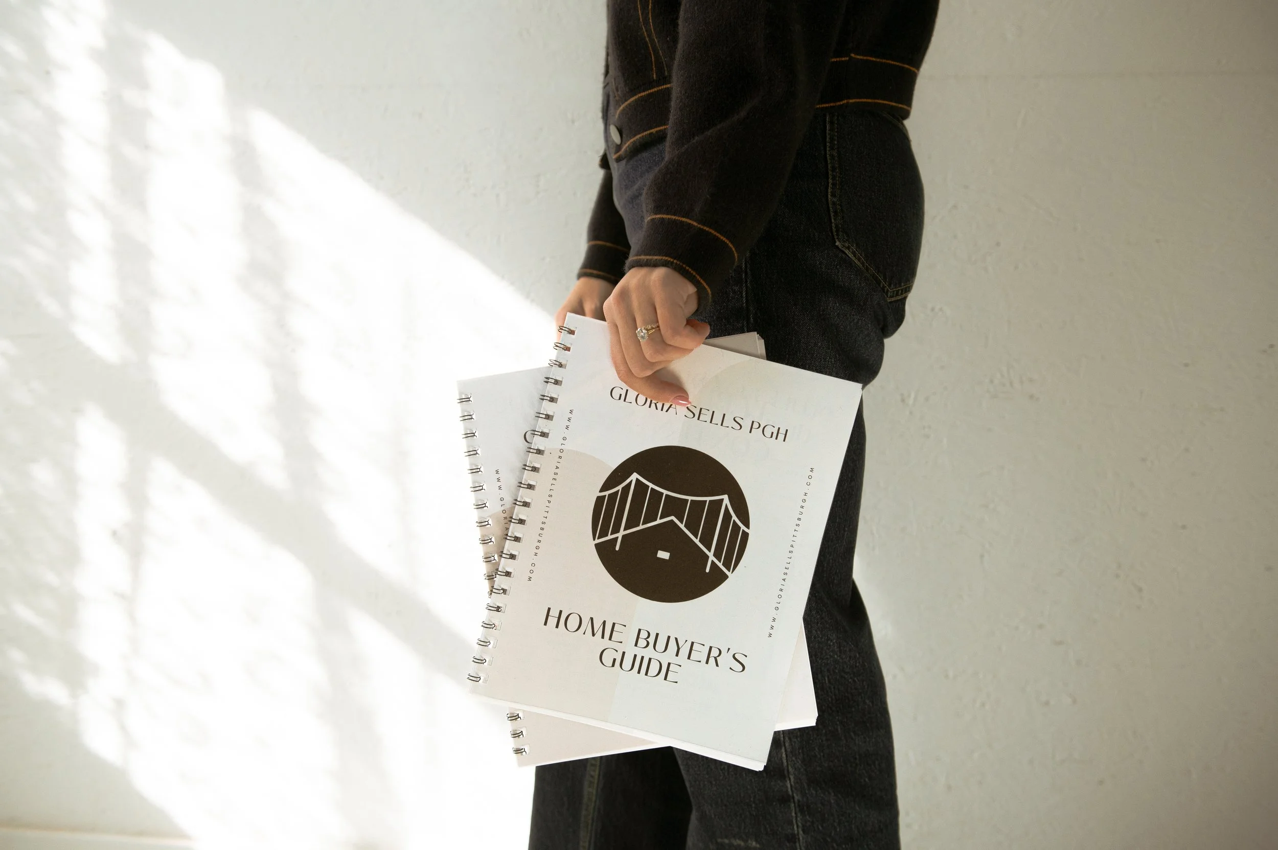 Person holding a home buyer's guide and property information booklet, wearing black jeans and a black jacket with orange stitching, against a plain light-colored wall.