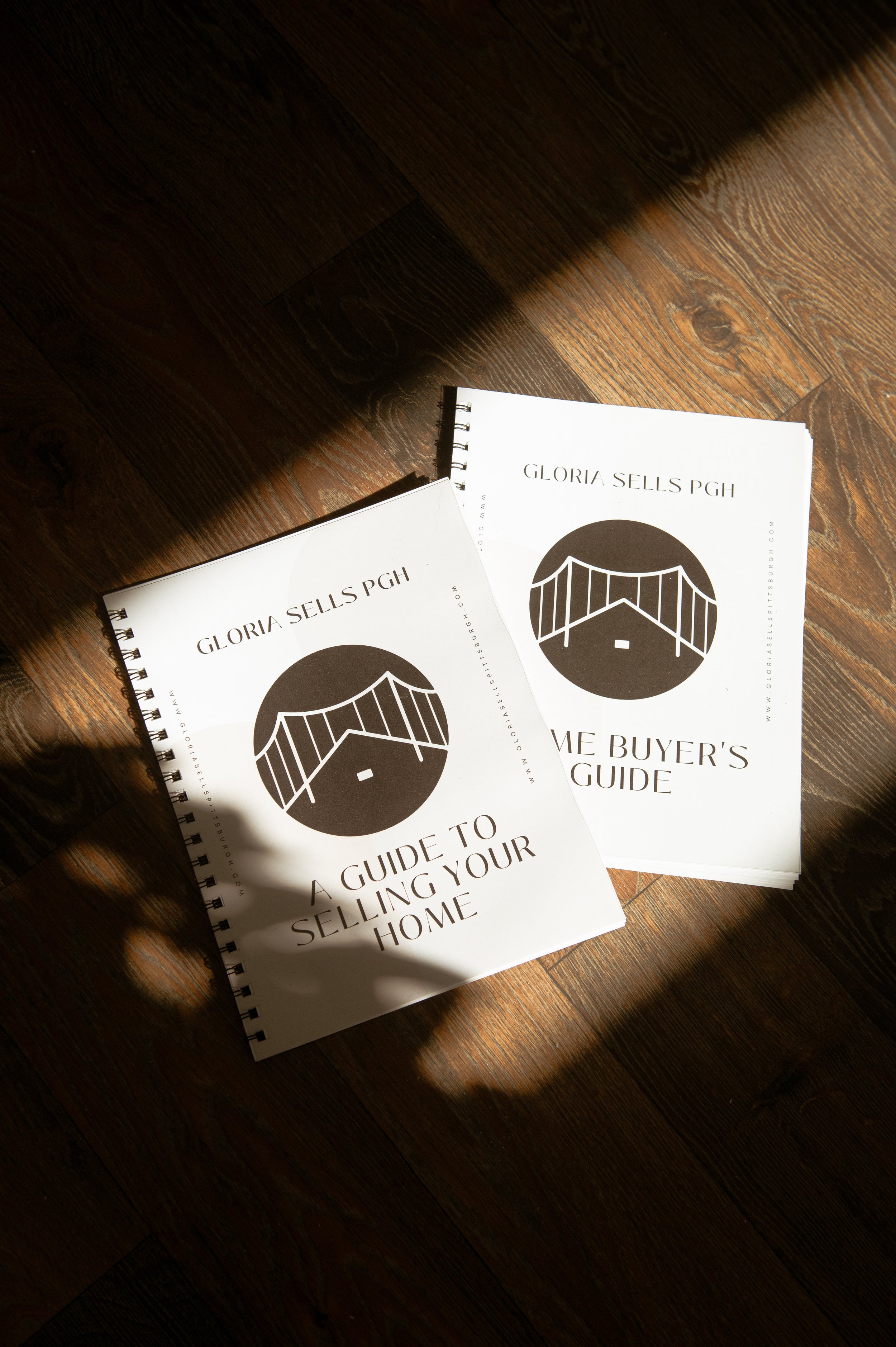 Two white spiral-bound booklets titled "Guide to Selling Your Home" and "Gloria Sells PGH" placed on a wooden floor with sunlight and shadow patterns.